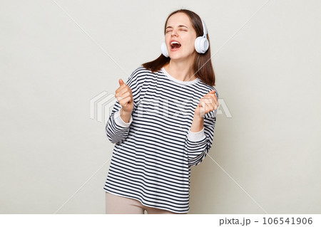 Overjoyed young woman wearing striped shirt listening to music in headphones isolated over gray background dancing and singing favorite song. 106541906
