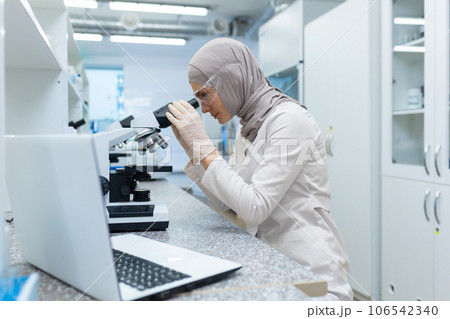 Young Muslim woman in hijab. a scientist, a pharmacist, a doctor works in a laboratory. Sits at the table and looks into the microscope, conducts research and analysis. 106542340