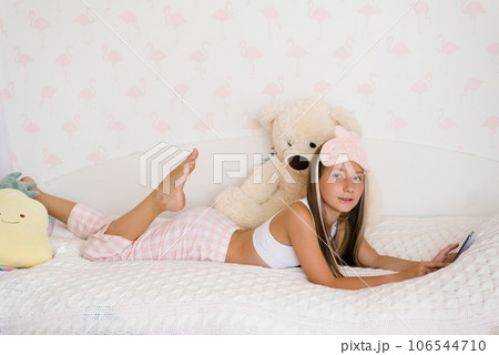 Beautiful teenage girl in pajamas is lying on a cozy bed on her stomach and surfing on the phone in the house in the morning, the younger generation is addicted to gadgets 106544710