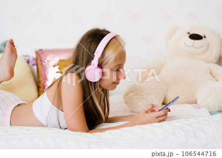Beautiful teenage girl is relaxing in bed using a touchscreen smartphone, listening to music with a wireless headset, surfing the Internet in the bedroom, relaxing comfortably, smiling, enjoying a 106544716