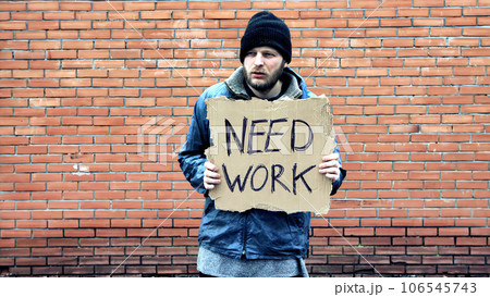 Homeless holds piece of cardboard with inscription Need Work. Refugee is at brick wall looking for job. Male tramp in dirty clothes. Below poverty line. Bearded fugitive beggar. Immigrant. Drunkenness 106545743