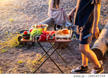 A man prepares a vegetable salad in nature, fresh peppers and tomatoes on the table. Rest in the company of friends in the summer in nature. Camping in the forest. Sandy bank of the river. 106545744