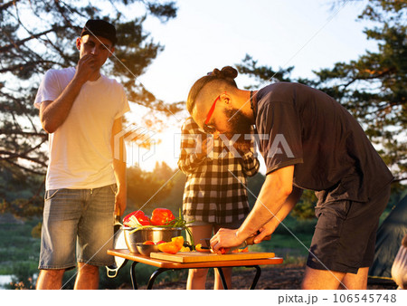 Stylish man with a beard and black glasses cuts fresh vegetables peppers and tomatoes to prepare a salad in nature. Vacation with friends in nature. Tourism as a hobby. Evening sunset. Stylish man with a beard and black glasses cuts fresh vegetables peppers and tomatoes to prepare a salad in nature. Vacation with friends in nature. Tourism as a hobby. Evening sunset. 106545748