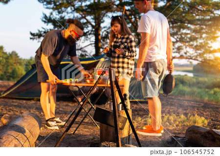 A young man with a beard and black glasses is preparing a salad of vegetables in nature in the company of friends. Outdoor recreation in summer. Tourism, evening sunset, lifestyle A young man with a beard and black glasses is preparing a salad of vegetables in nature in the company of friends. Outdoor recreation in summer. Tourism, evening sunset, lifestyle 106545752