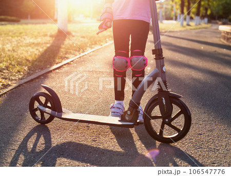 Legs of a girl in protective equipment against injuries on the background with a scooter. Safety in dangerous sports, close-up. Sunset Legs of a girl in protective equipment against injuries on the background with a scooter. Safety in dangerous sports, close-up. Sunset 106547776