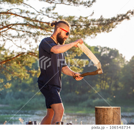 A handsome bearded man in black glasses is chopping firewood in nature in the summer against the background of a sunset. Outing for a picnic. Camping. 106548734