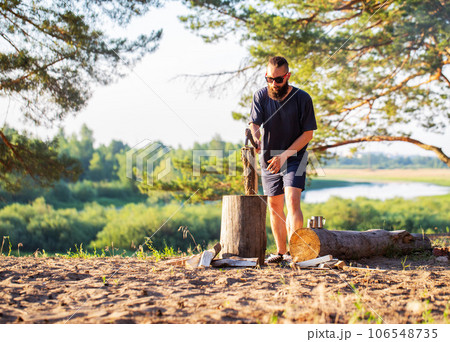A man with a beard, a lumberjack with an ax in his hands, cuts wood in nature in the summer against the backdrop of a forest and a river bank. Camping. Survival, travel A man with a beard, a lumberjack with an ax in his hands, cuts wood in nature in the summer against the backdrop of a forest and a river bank. Camping. Survival, travel 106548735