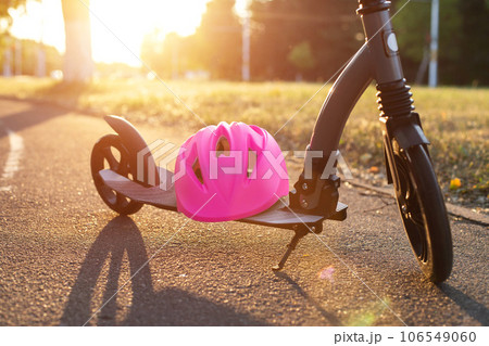 A pink helmet lies on a children's scooter against the backdrop of a sunset in the summer. The concept of safety while riding a scooter, close-up 106549060