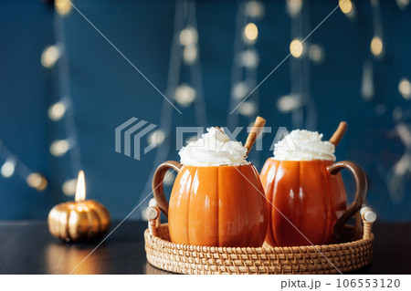 Pumpkin shaped cups of hot drink with whipped cream, cinnamon stick on black table with dark background. Pumpkin latte with spices. Cozy autumn beverage for Thanksgiving, Halloween holidays at home. Pumpkin shaped cups of hot drink with whipped cream, cinnamon stick on black table with dark background. Pumpkin latte with spices. Cozy autumn beverage for Thanksgiving, Halloween holidays at home. 106553120