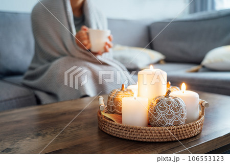 Focus on autumn fall cozy mood decor composition. Orange pumpkins, burning candles with a blurred woman covered with plaid and drinking tea on the sofa. Movie night party at home. Cozy relaxing time. 106553123