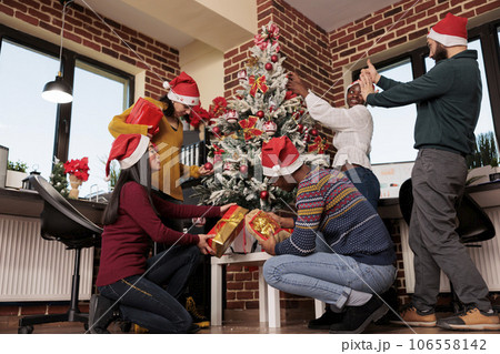 Young startup company employees sharing new year gifts and celebrating holiday in decorated office. Diverse men and women coworkers exchanging presents at corporate christmas party 106558142