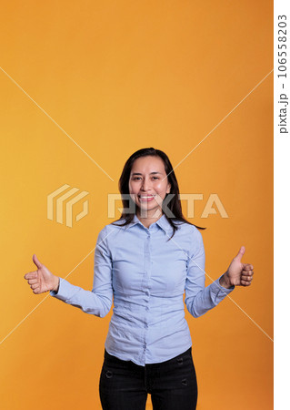 Smiling asian woman doing thumbs up gesture in front of camera showing approval and agreement gesture with fingers in studio. Satisfied model gesturing okay and positive symbol over yellow background 106558203