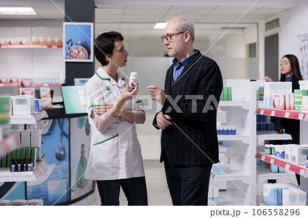 Elderly man in glasses buying prescripted heart medication in drugstore, choosing tablets for cardiac disease treatment. Medical worker holding medicine bottle and explaining side effects to customer Elderly man in glasses buying prescripted heart medication in drugstore, choosing tablets for cardiac disease treatment. Medical worker holding medicine bottle and explaining side effects to customer 106558296