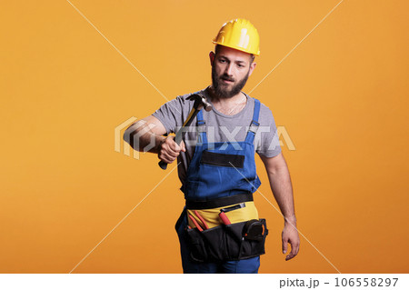 Construction worker using hammer on renovation project, standing over yellow background. Professional male renovator doing reconstruction work with sledgehammer and tools, studio shot. 106558297