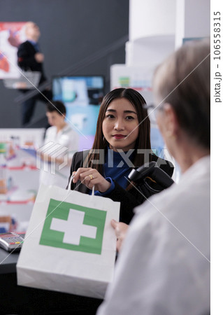 Woman buying pills at pharmacy cash register desk and listening to drugstore cashier. Pharmaceutical worker giving medical products shopping bag to young asian customer at checkout Woman buying pills at pharmacy cash register desk and listening to drugstore cashier. Pharmaceutical worker giving medical products shopping bag to young asian customer at checkout 106558315