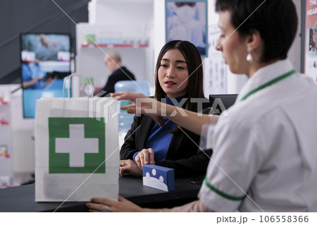 Pharmacy store customer buying vitamin and paying at drugstore checkout. Pharmaceutical assistant selling supplement to young asian client and putting multivitamin package in package Pharmacy store customer buying vitamin and paying at drugstore checkout. Pharmaceutical assistant selling supplement to young asian client and putting multivitamin package in package 106558366