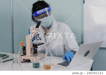 Indian Scientist man looking Microscope using laptop analyze research data in science laboratory. Asian biochemistry scientists wear white lab coat, mask face shield use microscope in laboratory tech 106561121