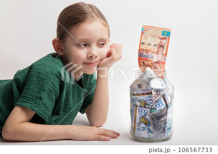 Kid with a large transparent jar with Kazakhstani tenge money on a white background 106567733
