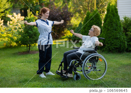 A nurse is exercising with an elderly patient in a wheelchair outdoors. 106569645