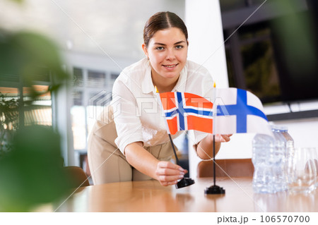 Positive young woman putting flag of Norway on the table with flag of Finland in conference room 106570700