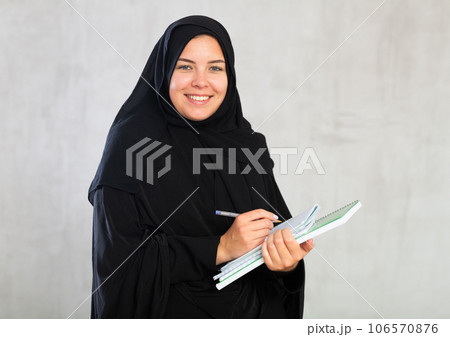 gleeful girl in hijab holds several notebooks and writes important note in her diary 106570876