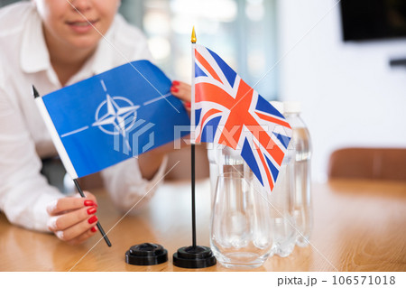 Little flag of Great Britain on table and flag of NATO put next to it by young woman Little flag of Great Britain on table and flag of NATO put next to it by young woman 106571018