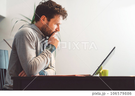 Side view waist up bearded caucasian man sitting by the table looking at his laptop, online studying from home Side view waist up bearded caucasian man sitting by the table looking at his laptop, online studying from home 106579886