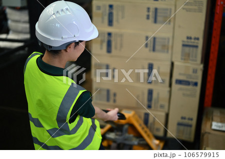 Strong man worker wearing hardhat and vests pulling pallet truck walking through rows of storage racks with merchandise 106579915