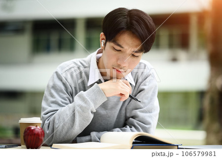 Concentrated asian male student reading book, preparing for exam or, doing homework remotely in the park 106579981