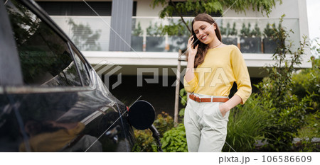Close up of beautiful woman calling while charging her electric car on the street. Close up of beautiful woman calling while charging her electric car on the street. 106586609