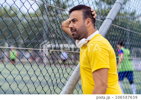 Worried man with wireless earphones watching football club training on large field leaning on metal fence grid fan looking at players actions on public arena 106587639