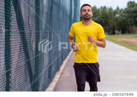 Man with beard wearing comfortable tracksuit running along road past high mesh fence on sports ground in urban park activities for health and body development 106587646