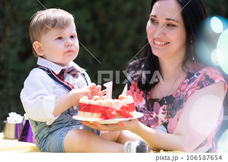delightful baby boy, accompanied by his mother, enjoys his birthday celebration featuring a cake topped with a single candle. soap bubbles fly delightful baby boy, accompanied by his mother, enjoys his birthday celebration featuring a cake topped with a single candle. soap bubbles fly 106587654