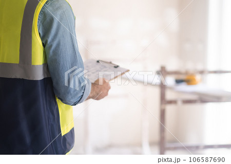 Professional engineer holding clipboard with plan of house interior renovation in hands standing against shabby wall and construction equipment in light room 106587690