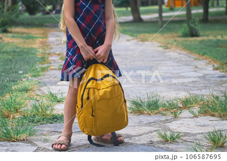 Schoolgirl in colorful dress stands on tiled path in autumn park holding big yellow backpack girl with long blonde hair goes to school in sunny morning Schoolgirl in colorful dress stands on tiled path in autumn park holding big yellow backpack girl with long blonde hair goes to school in sunny morning 106587695