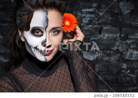 Pensive beautiful girl with skull make-up on face posing with flower indoors. Closeup portrait of female with decorative make-up placing gerbera over ear and smiling at camera. Halloween concept. Pensive beautiful girl with skull make-up on face posing with flower indoors. Closeup portrait of female with decorative make-up placing gerbera over ear and smiling at camera. Halloween concept. 106588552