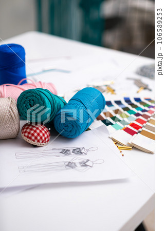 Macrame threads coils and needle cushion on tailor table in fashion studio colorful yarn sketch and swatch for dress craft in sewing workshop seamstress supplies closeup Macrame threads coils and needle cushion on tailor table in fashion studio colorful yarn sketch and swatch for dress craft in sewing workshop seamstress supplies closeup 106589253