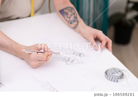 Lady designer draws clothes model on paper at table in fashion atelier tailor makes pencil sketch of apparel for women in sewing studio closeup dressmaking industry Lady designer draws clothes model on paper at table in fashion atelier tailor makes pencil sketch of apparel for women in sewing studio closeup dressmaking industry 106589281