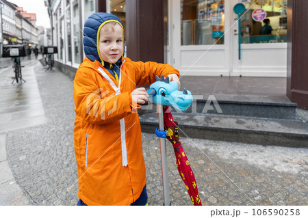 Cute little lonely caucasian kid boy in bright orange waterproof raincoat enjoy riding scooter walking on european city street outside. Autumn weather season outdoor sport activities 106590258
