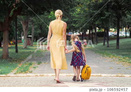 A lively schoolgirl, her yellow backpack bouncing, playfully hops while walking with her mother across the park on their way to school.  106592457