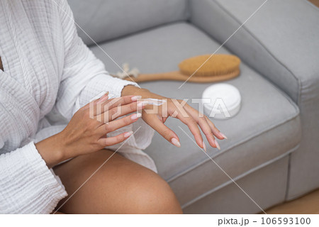 Home spa: A close-up reveals a woman, bathrobe-clad, joyfully applying moisturizing cream to her hand, savoring a luxurious body skincare ritual.  106593100