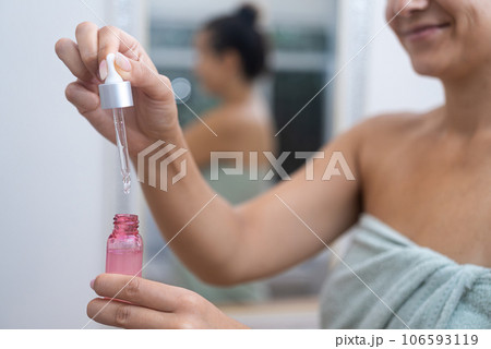 Smiling woman with towel dripping facial serum over small bottle in bathroom pretty lady showing hyaluronic acid used rejuvenating skincare at home closeup 106593119