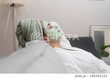 Sofa Sanctuary: Wrapped in her bathrobe, she relaxes on a comfortable sofa, embracing the tranquility of a facial moisturizing mask during her home spa session.  106593123