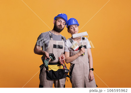 Exhausted team of contractors working with construction tools, falling asleep after renovating project. Man and woman builders holding power drill, painting brush or roller and pair of pliers. Exhausted team of contractors working with construction tools, falling asleep after renovating project. Man and woman builders holding power drill, painting brush or roller and pair of pliers. 106595962