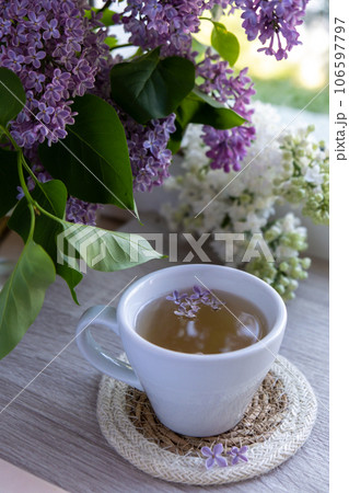 Tasty black tea in white cup on windowsill with aromatic lilac flowers. Spring composition Cup of lilac tea drinking recipe flowering branches of purple lilac. Still life for copy space greeting card Tasty black tea in white cup on windowsill with aromatic lilac flowers. Spring composition Cup of lilac tea drinking recipe flowering branches of purple lilac. Still life for copy space greeting card 106597797