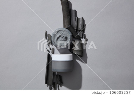 Unlabeled cosmetic jars filled with clay mask decorated with several activated charcoal sticks. Gray background. Activated charcoal has absorbent properties 106605257
