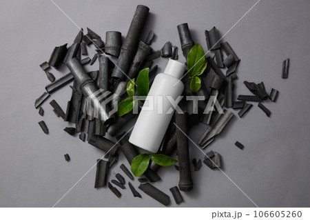 Blank label bottle placed on a pile of activated charcoal sticks. Few green tea leaves decorated on gray background. Mockup of skin care cosmetic bottle of beauty facial 106605260