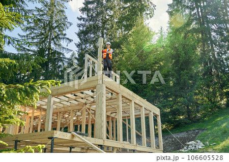 Front view of male worker bilding wooden house, standing high, holding wooden gilder, putting. Builder wearing helmet, constructing premises in forest. Concept of building. 106605528