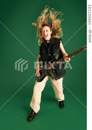 Full-length image of young girl in stylish clothes emotionally playing electric guitar against green studio background 106607283