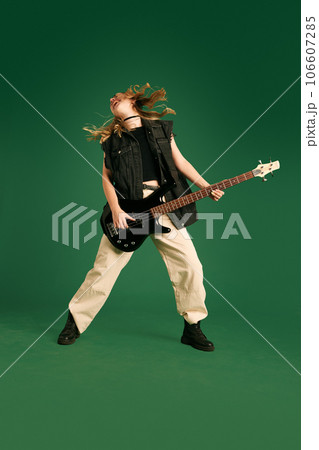 Full-length image of young girl in stylish clothes emotionally playing electric guitar against green studio background Full-length image of young girl in stylish clothes emotionally playing electric guitar against green studio background 106607285